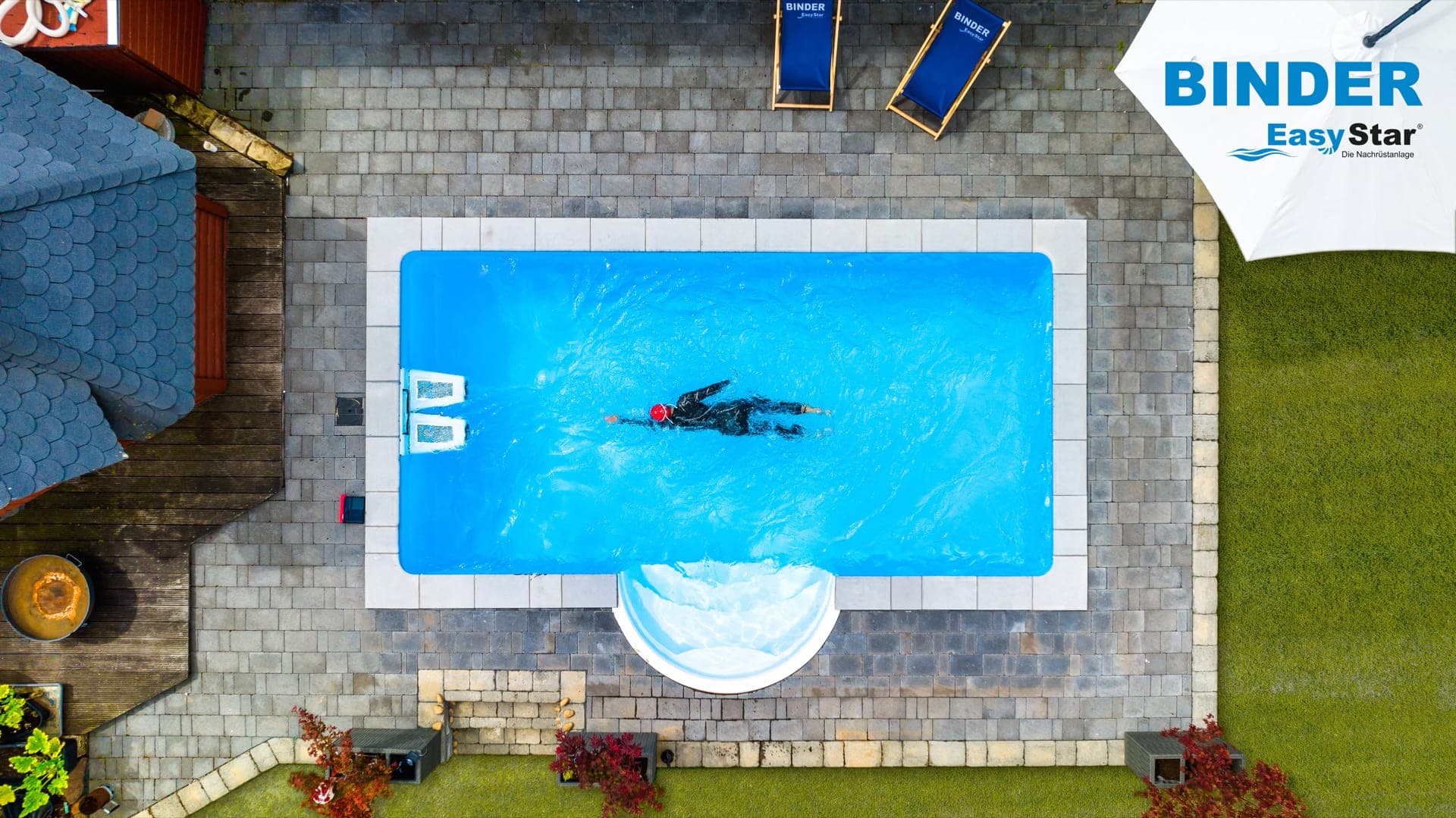 Aerial drone view of a residential villa pool with a swimmer, surrounded by stone terrace, sun loungers, parasol, and garden lawn